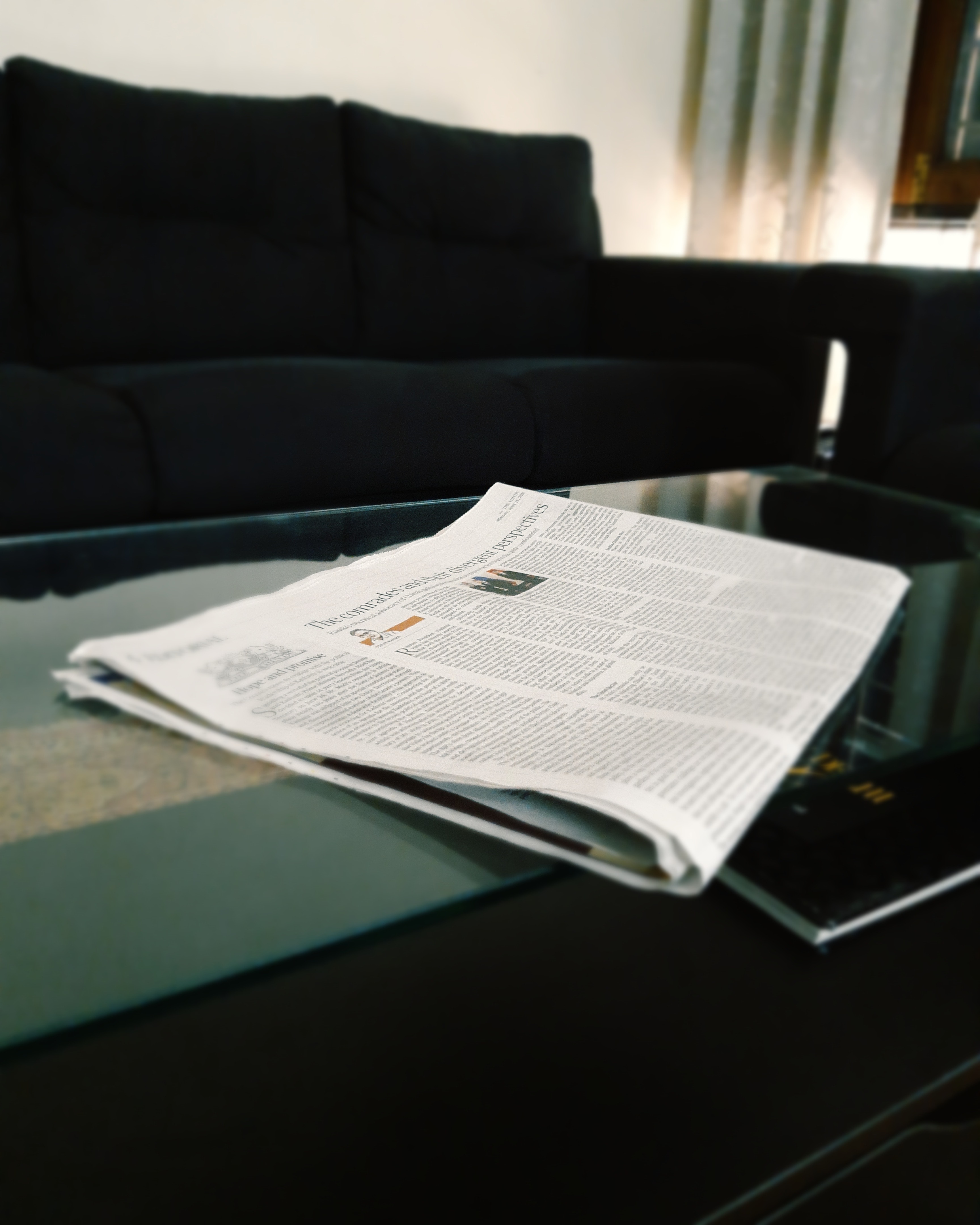 Eine Zeitung liegt auf einem Glastisch, im Hintergrund ist ein schwarzes Sofa zu sehen. (Quelle: Unsplash)